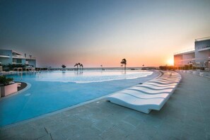 Piscina com borda curva, área de descanso com bancos brancos e vista para o oceano ao pôr do sol.