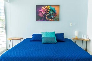 Quarto com cama, duas almofadas, mesa de cabeceira, abajur e um quadro de uma árvore na parede.