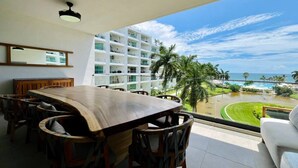 A dining area with a large wooden table and chairs, a white sofa, and a view of a hotel building and beach. - 406 Increible depa con jacuzzi priv y vista al mar (Nuevo Vallarta)
