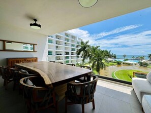 Ein Essbereich mit einem großen Holztisch und Stühlen, einem weißen Sofa und Blick auf ein Hotelgebäude und den Strand.