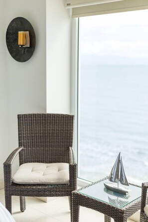 A wicker chair with a cushion, a glass table with a model ship, and a view of the sea through a large window. - Depto. Playa Vista Mar Villa Magna Nuevo Vallarta (Nuevo Nayarit)