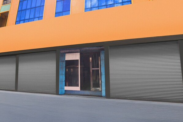 A building with a large orange facade, a central entrance with a glass door, and two closed roller shutters on either side.