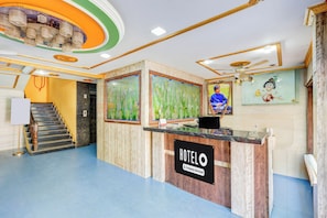 A hotel reception area with a marble counter, framed floral paintings, a staircase, and a ceiling fan.