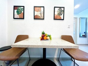 A dining area with a table and two chairs, a fruit bowl on the table, and three framed artworks on the wall. - Departamento/Estudio en Playa Royale, Nuevo Vta. (Nuevo Vallarta)