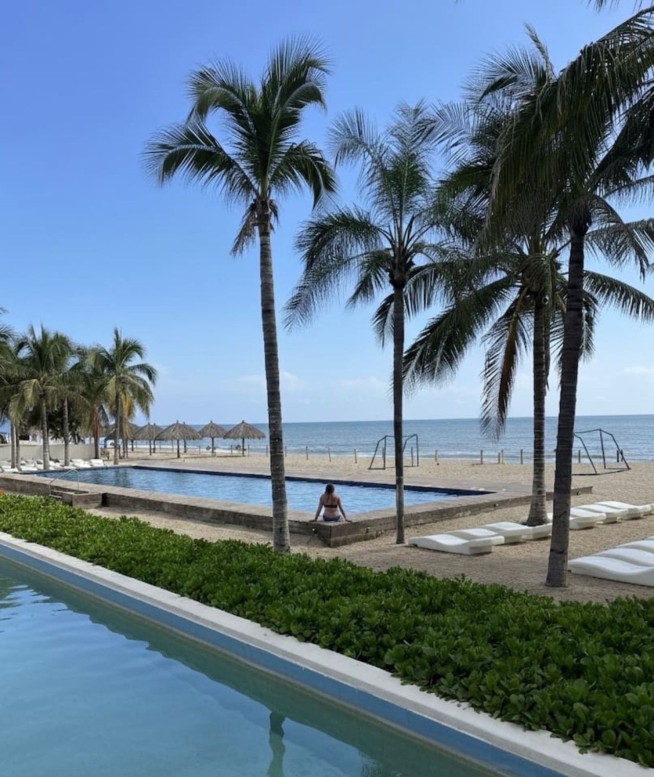 A pool area with palm trees, a beach, and a clear sky.