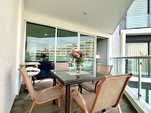 Uma varanda com mesa e cadeiras de vidro, um vaso com flores e vista para a cidade.