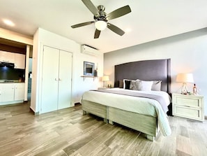 Quarto com cama, mesas de cabeceira, ventilador de teto e vista para a cozinha.