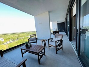 Varanda com vista para vegetação e uma estrada, mobiliada com cadeiras de madeira, uma mesa e uma mesa redonda pequena.