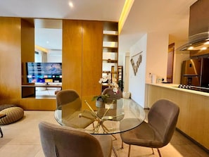 Uma sala de estar moderna com mesa de jantar de vidro, assentos confortáveis, TV e cozinha com coifa de aço inoxidável.