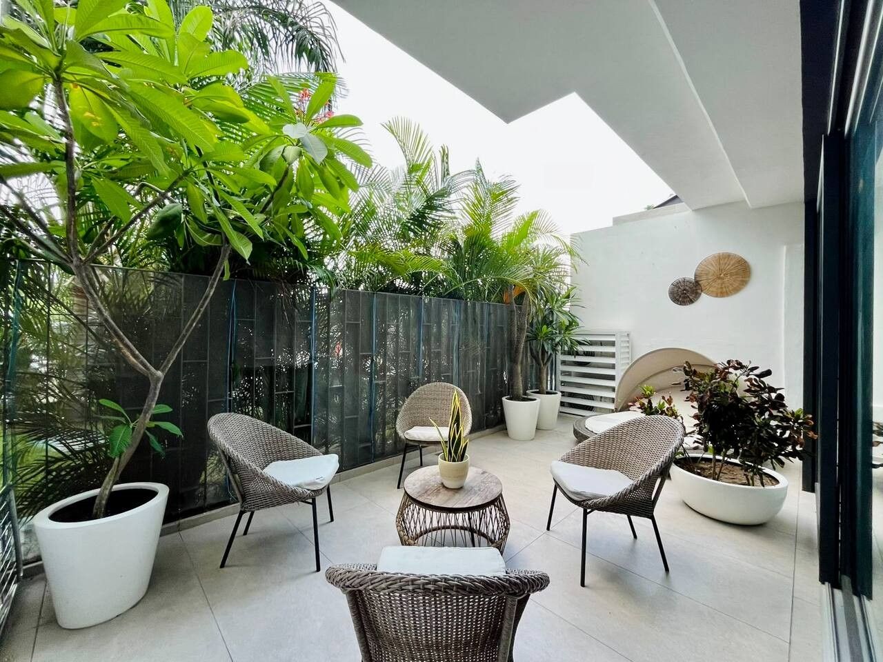 Un espace extérieur moderne, aménagé avec des chaises en osier et une table ronde, entouré de plantes en pot et délimité par une paroi en verre.