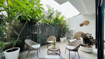 A modern outdoor seating area with wicker chairs and a round table, surrounded by potted plants and a glass wall.