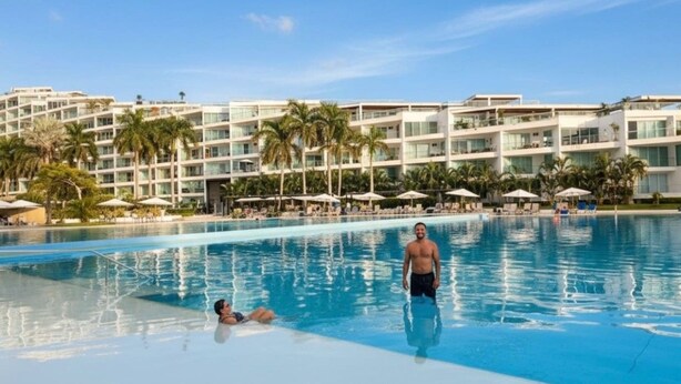 Una piscina alberghiera con superficie d'acqua cristallina e blu, circondata da palme e un edificio a più piani con balconi.