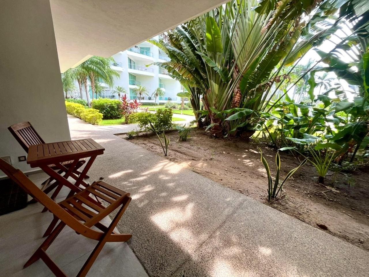 A covered outdoor area with a wooden table and chairs, surrounded by lush tropical plants and a view of a modern building in the background.