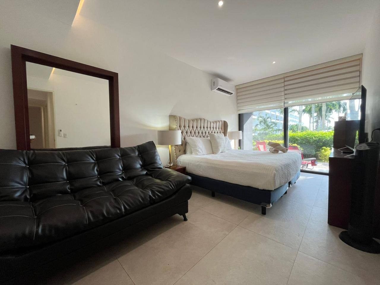 A modern bedroom with a large bed, a sofa, a TV, and a window with blinds.
