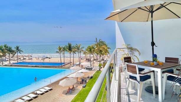 Un balcone con vista su una piscina e una spiaggia, un tavolo con sedie e un ombrellone grande.