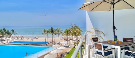 A balcony with a view of a pool and beach, a table with chairs, and a large umbrella.