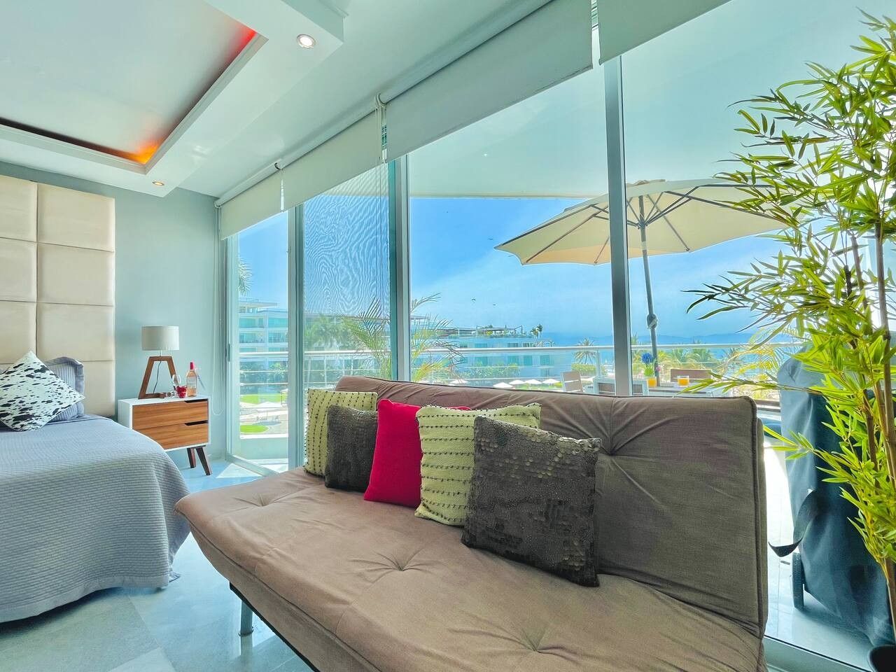 Quarto de hotel moderno com uma janela grande, um sofá com almofadas, uma mesinha de cabeceira e uma luminária.