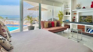 Un soggiorno moderno con un letto grande, un divano e una televisione. È presente un balcone con vista sull'oceano.