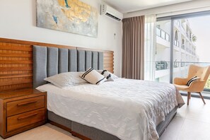 A hotel room with a large bed, a wooden nightstand, a chair, and a balcony with a view of a building. - Wake Up to Ocean Views – Aria Nuevo Vallarta (Nuevo Vallarta)