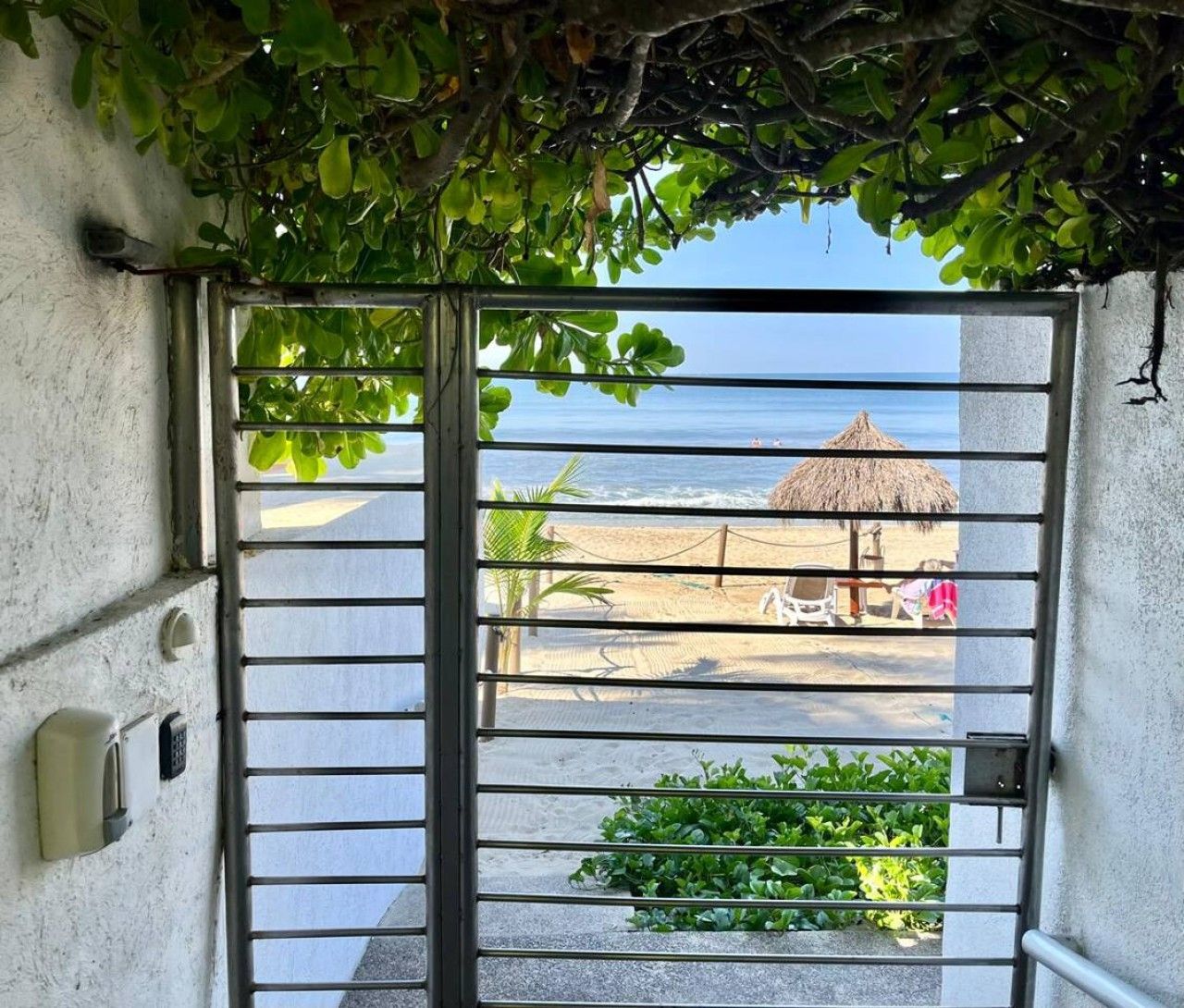 A metal gate with a view of a beach, a thatched umbrella, and a chair.