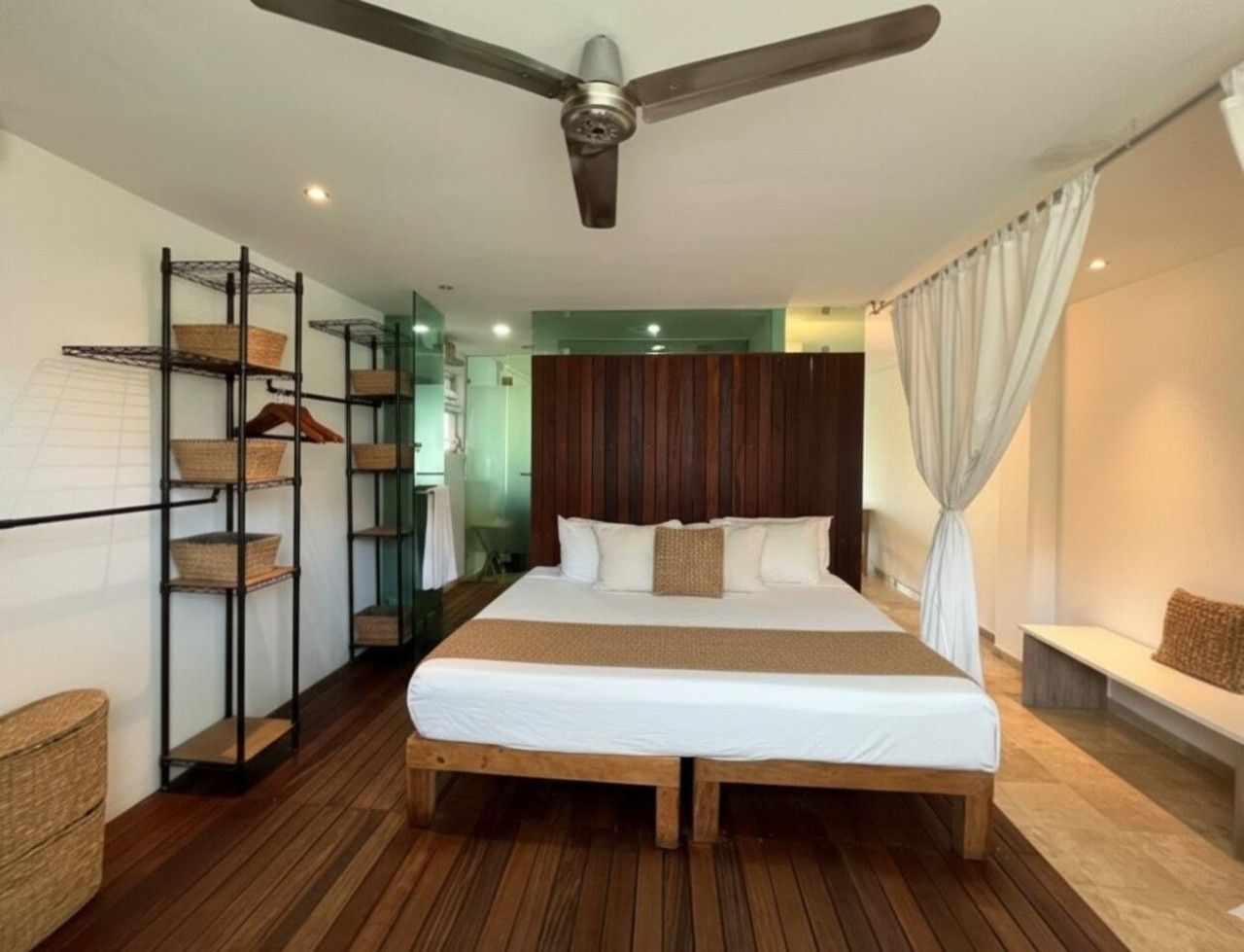 A hotel room with a large bed, a ceiling fan, a wooden floor, and a wall-mounted shelf with baskets.