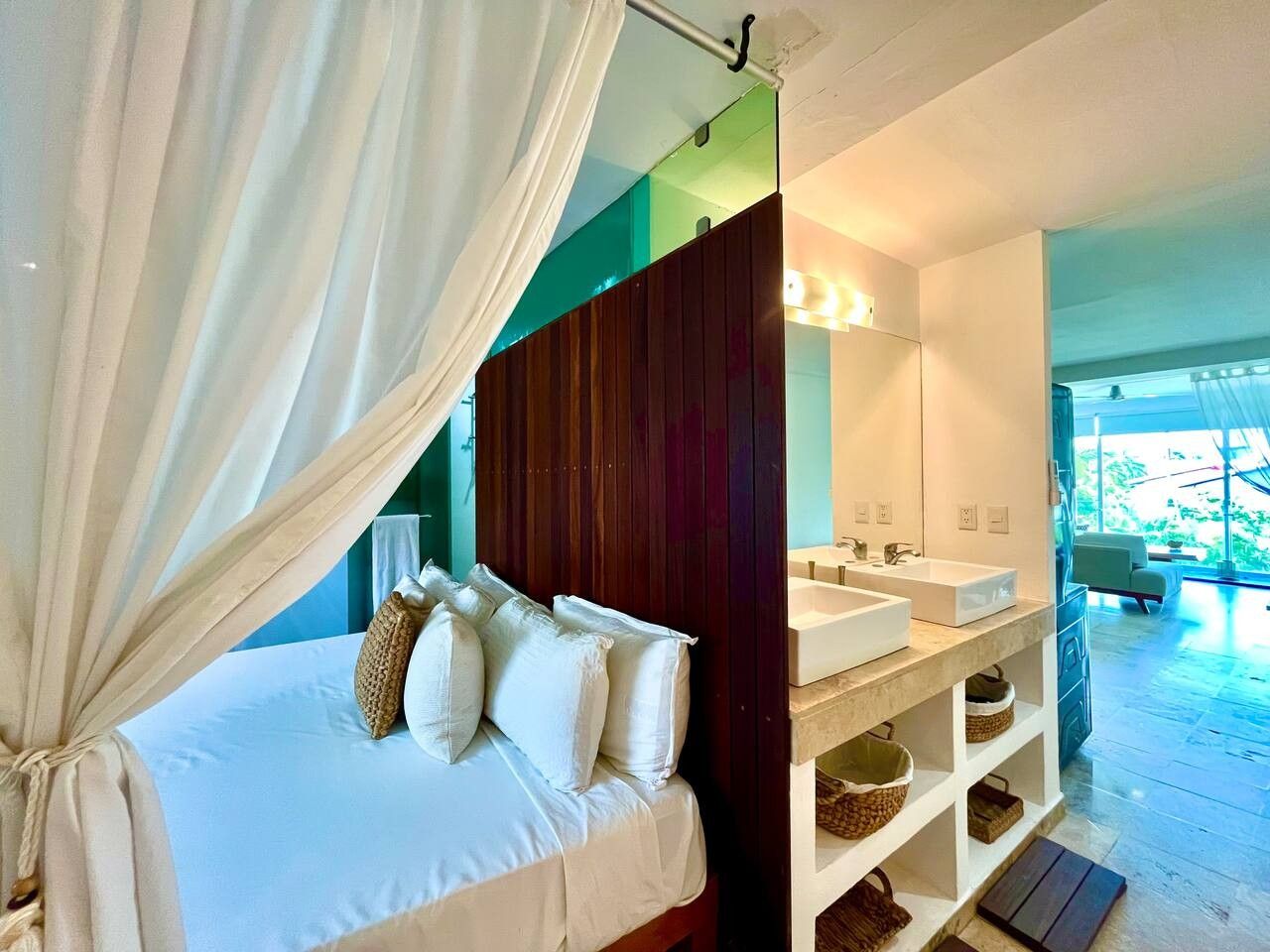 A bedroom with a bed, a bedside table, a mirror, and a bathroom with a sink and a shower.