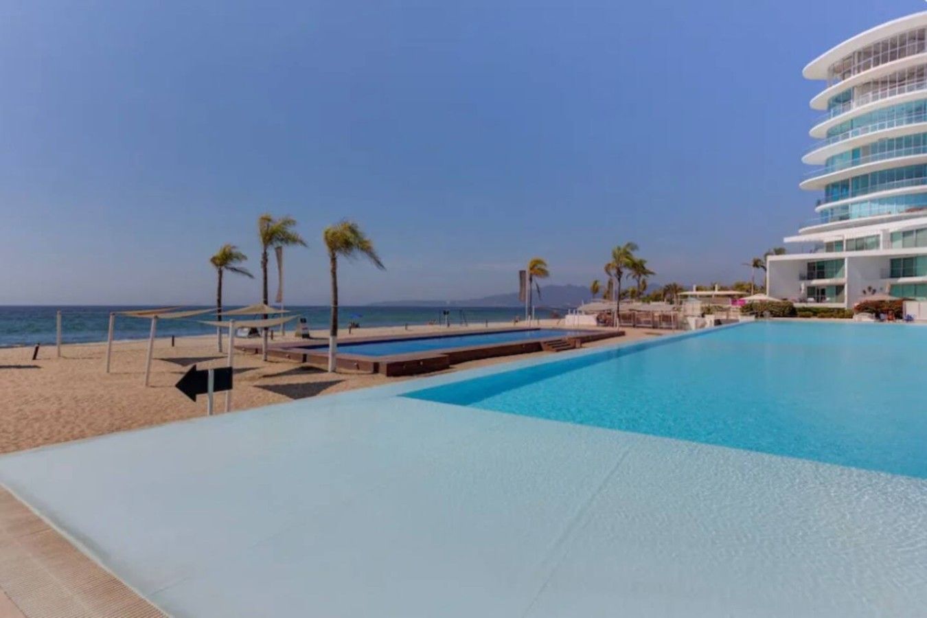 A swimming pool with a view of the beach and ocean, surrounded by palm trees and a modern building.