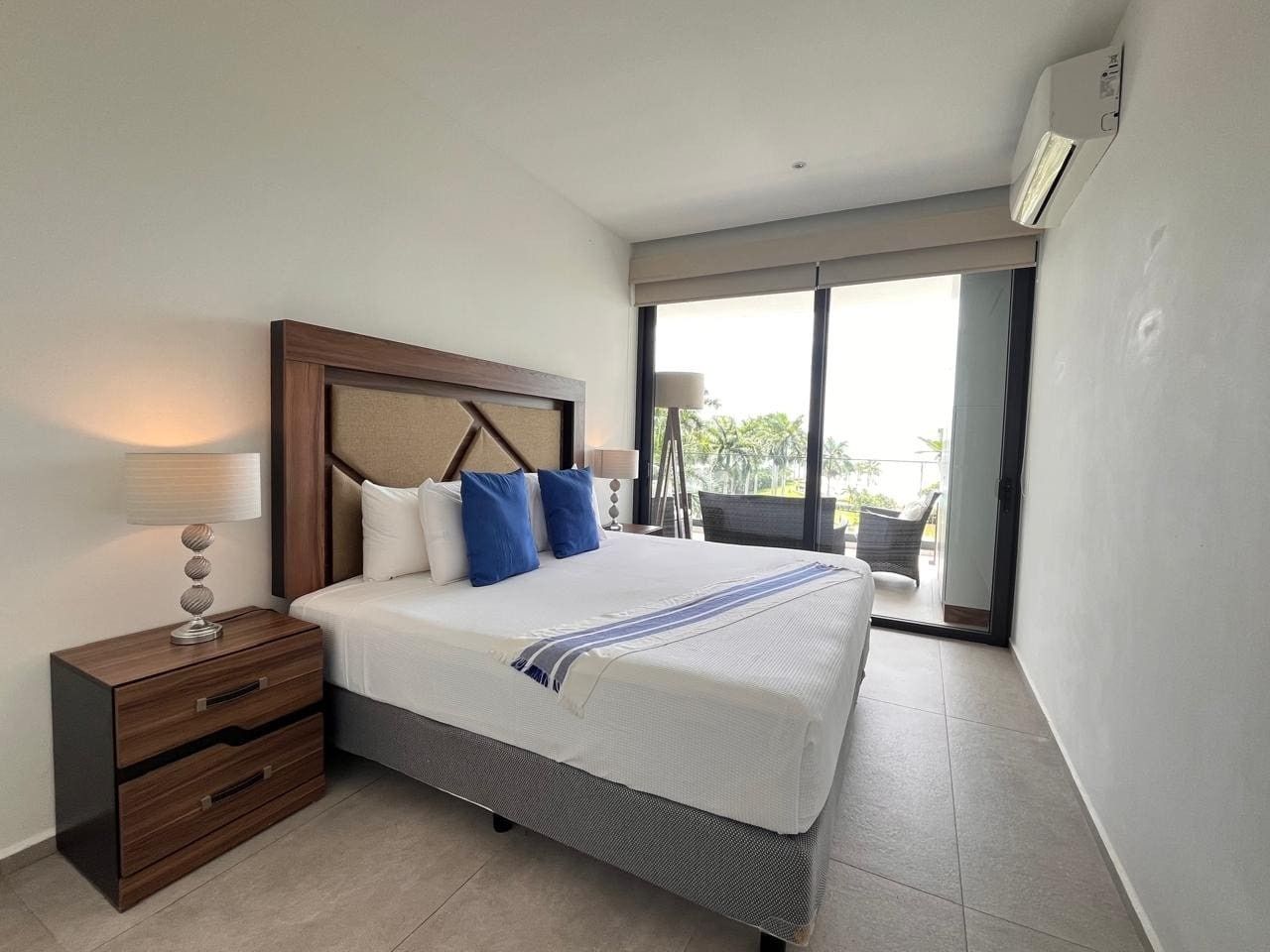 A hotel room with a bed, bedside table, lamp, and a view of the outdoors.