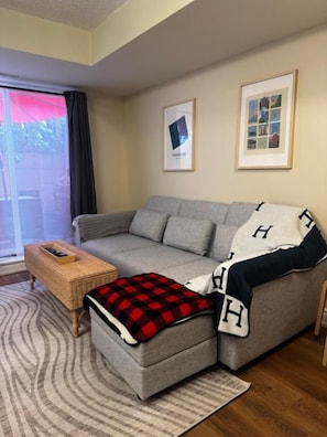 A living room with a grey sofa, a small ottoman, a wicker side table, and a framed picture on the wall. - DT Townhouse Room Steps to Transit (Toronto)