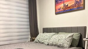A single bed with a grey headboard, a grey blanket, and a folded towel. There is a window with a closed blind and a curtain. A cityscape painting is on the wall above the bed.