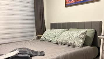 Un lit simple avec une tête de lit grise, une couverture grise et une serviette pliée. Il y a une fenêtre avec un store fermé et un rideau. Un tableau représentant un paysage urbain est accroché au mur au-dessus du lit.