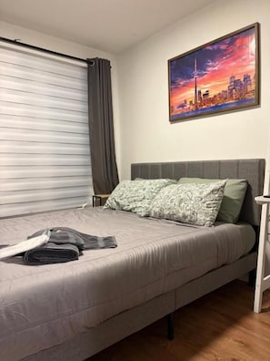 A single bed with a grey headboard, a grey blanket, and a folded towel. There is a window with a closed blind and a curtain. A cityscape painting is on the wall above the bed. - DT Townhouse Room Steps to Transit (Toronto)