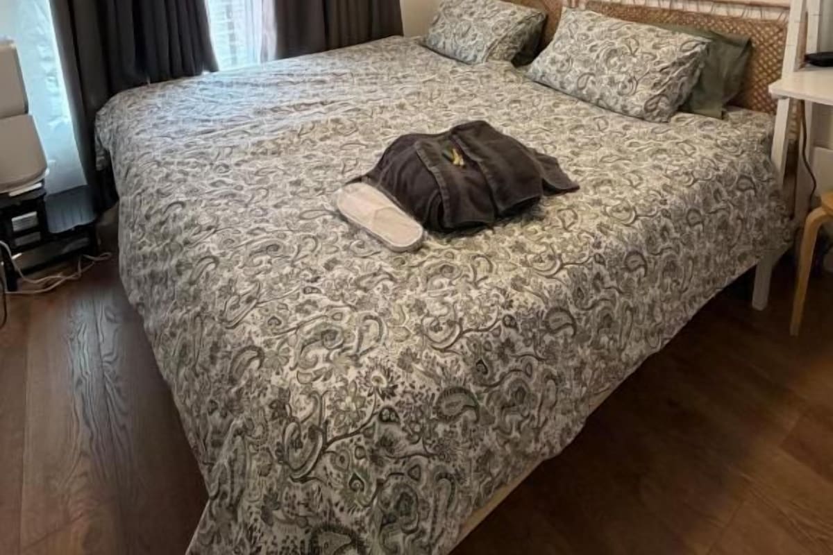 A bedroom with a double bed, a nightstand, a lamp, a painting, and a window with curtains.