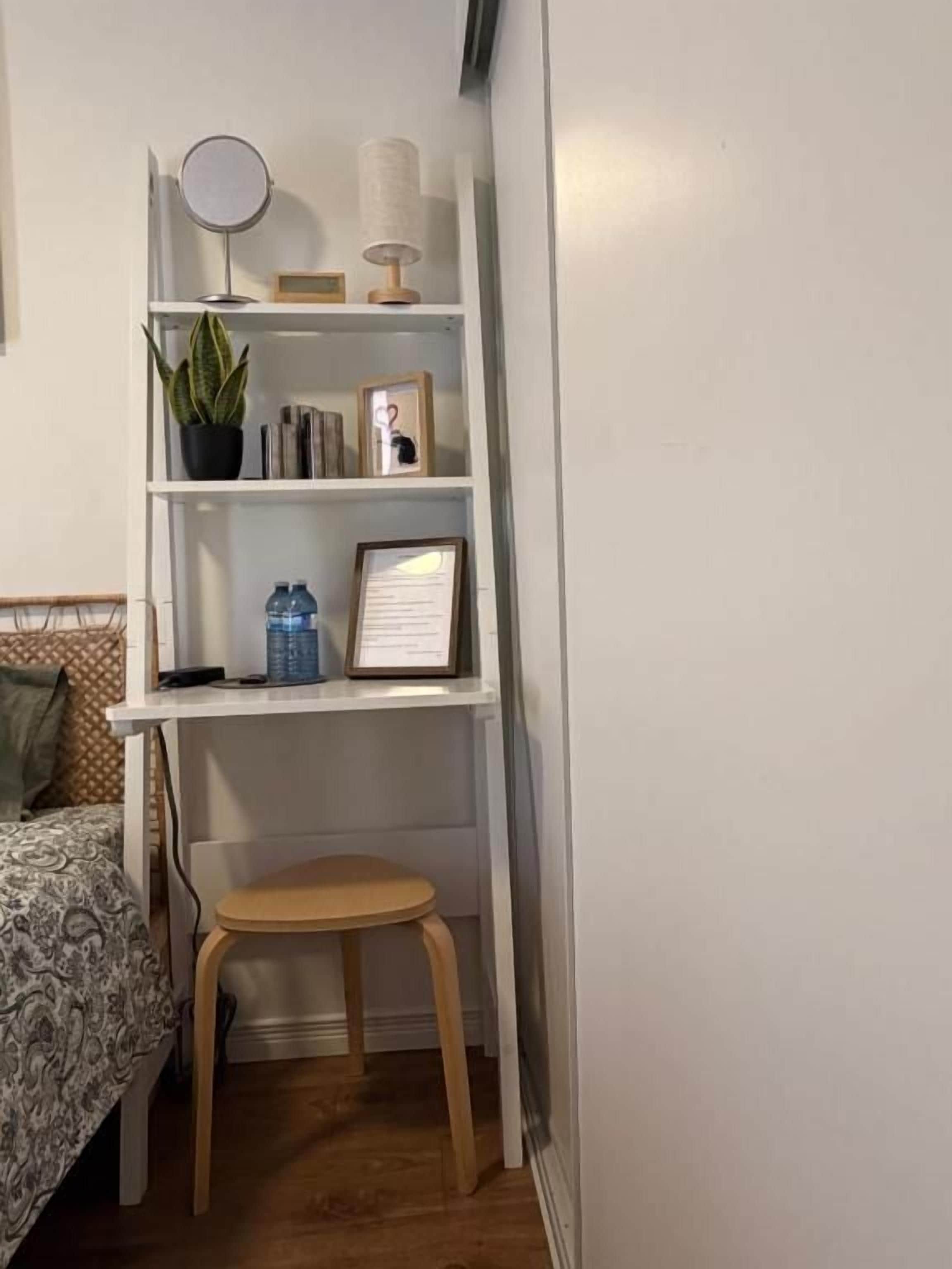 A small, neatly organized room with a bed, a wooden stool, a white shelf with various items, and a wall-mounted mirror.