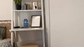 A small, neatly organized room with a bed, a wooden stool, a white shelf with various items, and a wall-mounted mirror.