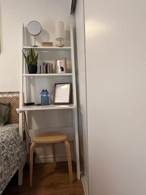 A small, neatly organized room with a bed, a wooden stool, a white shelf with various items, and a wall-mounted mirror.