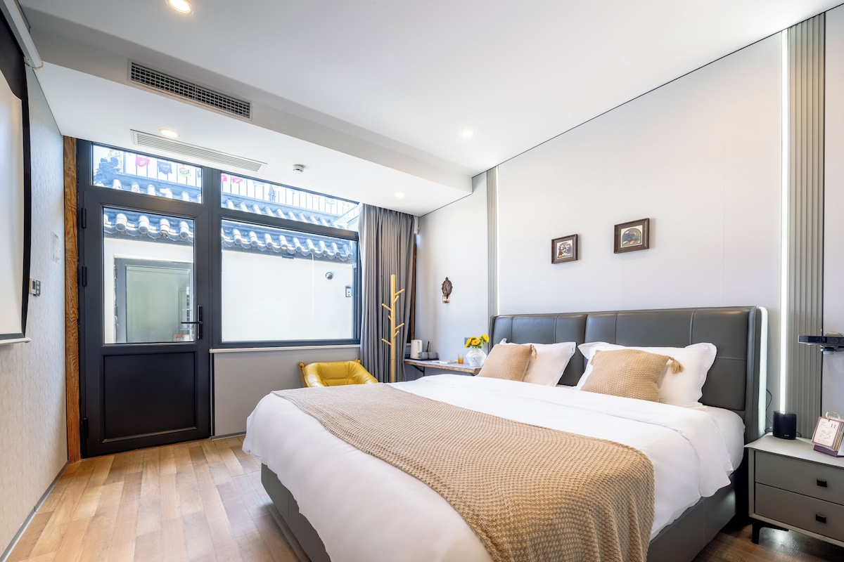 A modern hotel room with a large bed, a bedside table, a nightstand, a window with curtains, and a view of the outside.