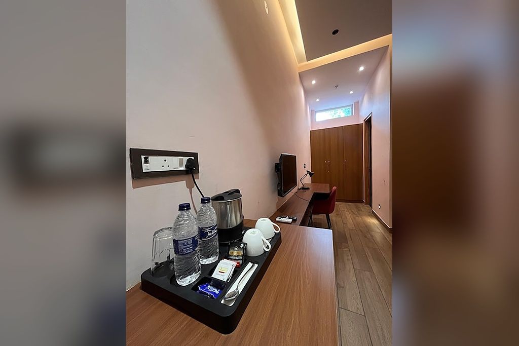 A hotel room with a wooden floor, a desk with a tray of amenities including water bottles, a kettle, and a cup, and a television on the wall.