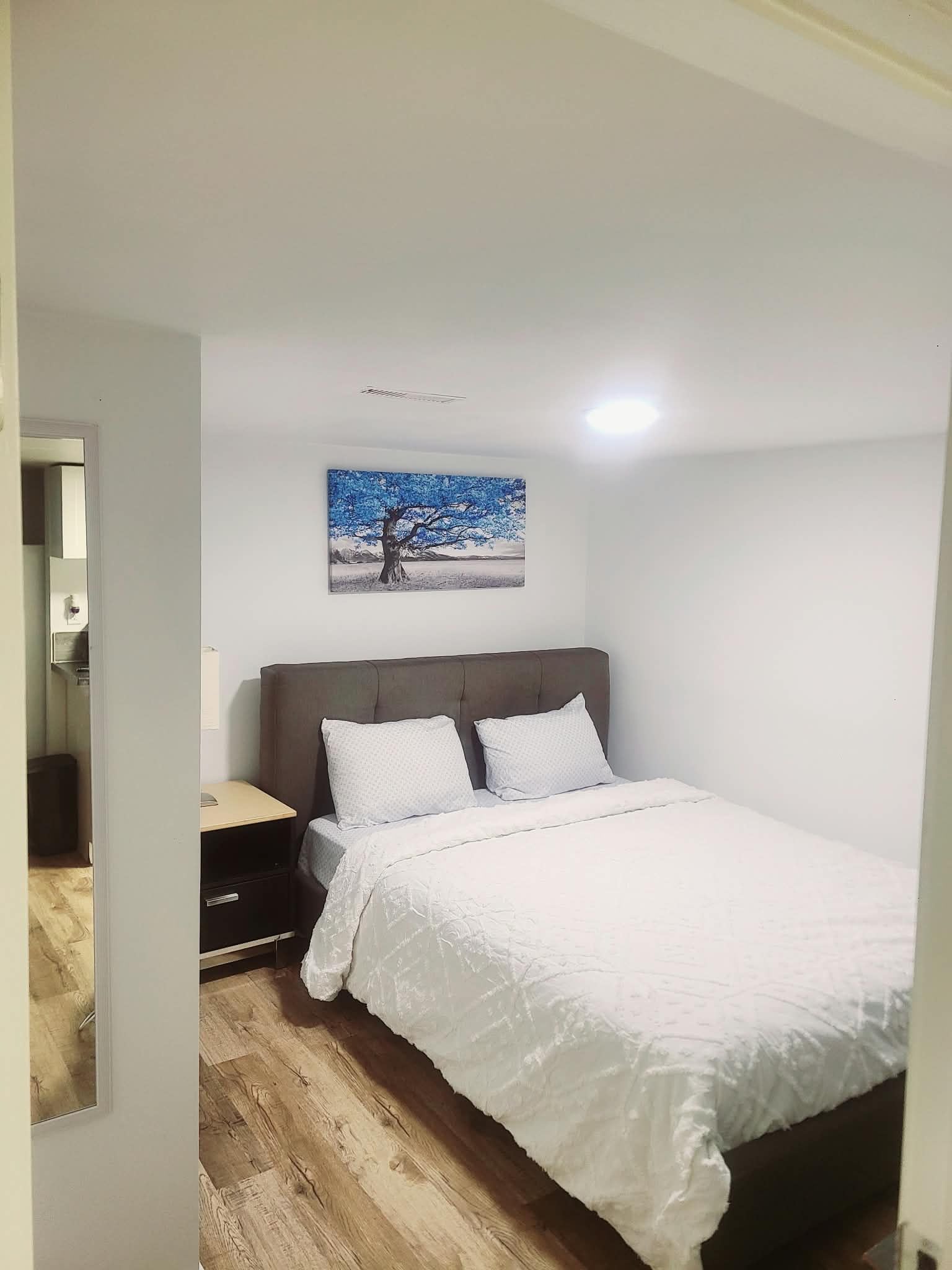 Une chambre à coucher avec un lit, une table de chevet, un tiroir, un tableau accroché au mur et un luminaire au plafond.