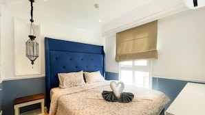 Un dormitorio con cabecera azul, cama con disposición de almohadas en forma de corazón, mesita de noche y ventana con persiana.