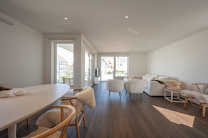 A spacious living area with a wooden floor, a sofa set, a dining table with chairs, and a view through large windows. - Elegant Apartment Near Casino (Knokke-Heist)