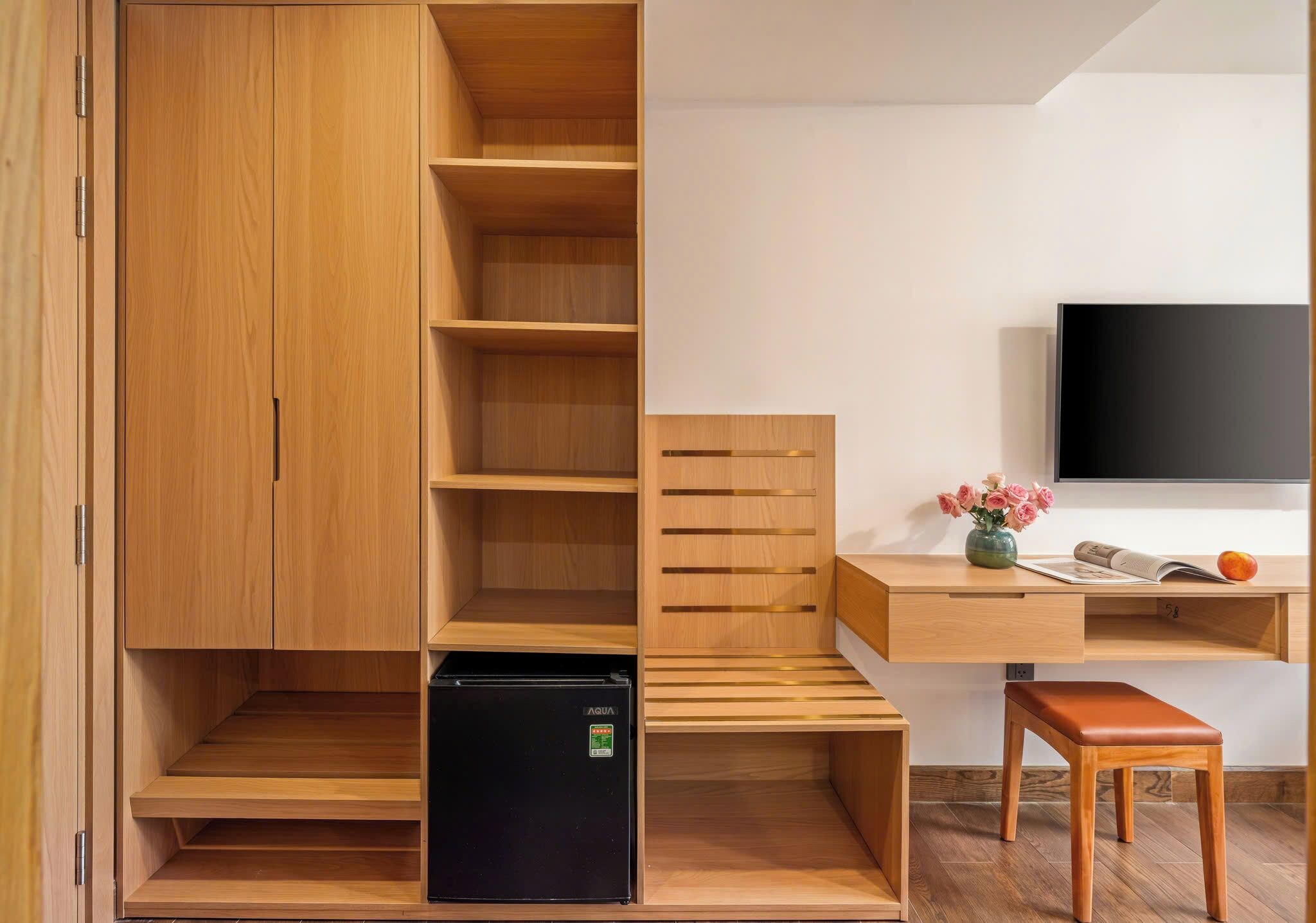 A compact room with a wooden wardrobe, a small black refrigerator, a wooden desk with a chair, and a flat-screen TV.
