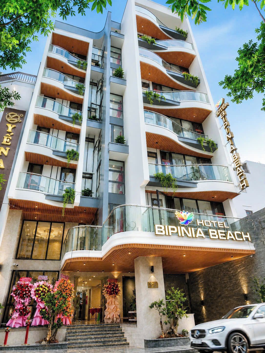 Et hotel med flere etager, altaner, en markant indgang og et skilt med teksten 'BIPINIA BEACH HOTEL'.