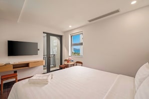 A hotel room with a bed, a desk with a flat-screen TV, a window with a view, and a door leading to a balcony. - Bipinia Beach Hotel (Quy Nhơn Nam)