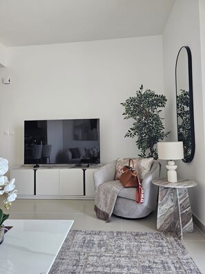 A modern living room with a flat-screen TV, a sofa, a coffee table, a side table with a lamp, a potted plant, and a large mirror.