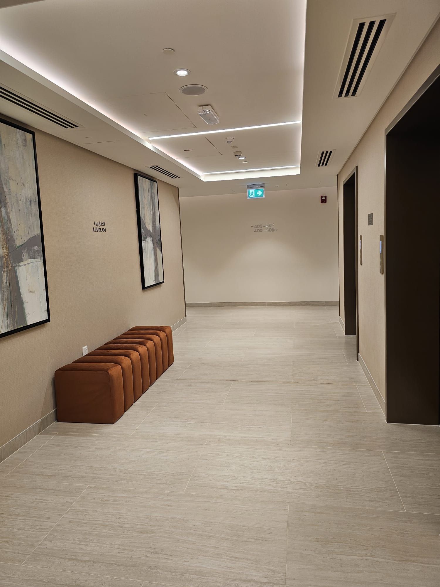 A hallway with a bench, ceiling lights, and emergency exit signs.