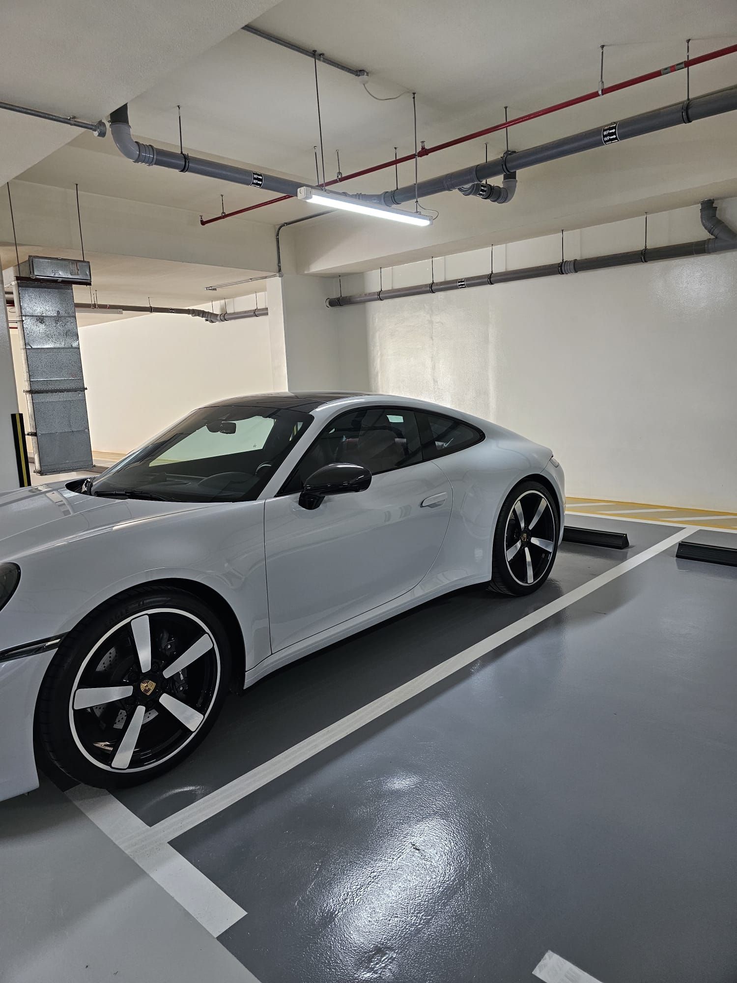 A white sports car parked in a clean, well-lit indoor parking garage with overhead pipes and a concrete floor.