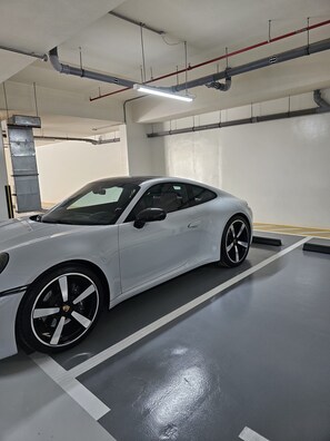 A white sports car parked in a clean, well-lit indoor parking garage with overhead pipes and a concrete floor.