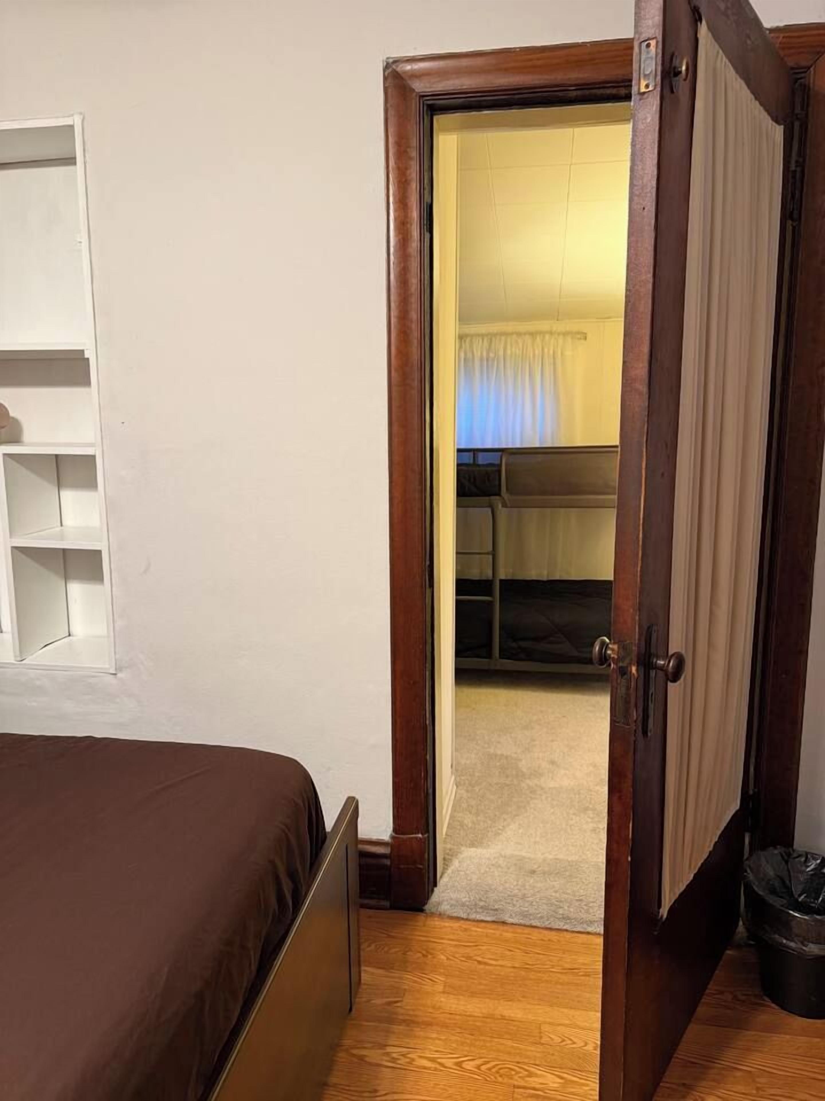 Ein Schlafzimmer mit einem Bett, einer Holztür, einem weißen Regal und Blick in einen angrenzenden Raum mit Bett und Vorhängen.