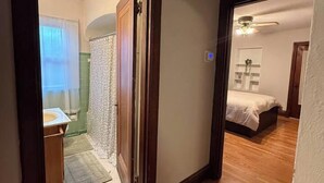 A hallway leading to a bedroom with a bed, a bathroom with a shower, and a wooden floor. - Historic STL Home near Wash U/Massive Backyard (University City)